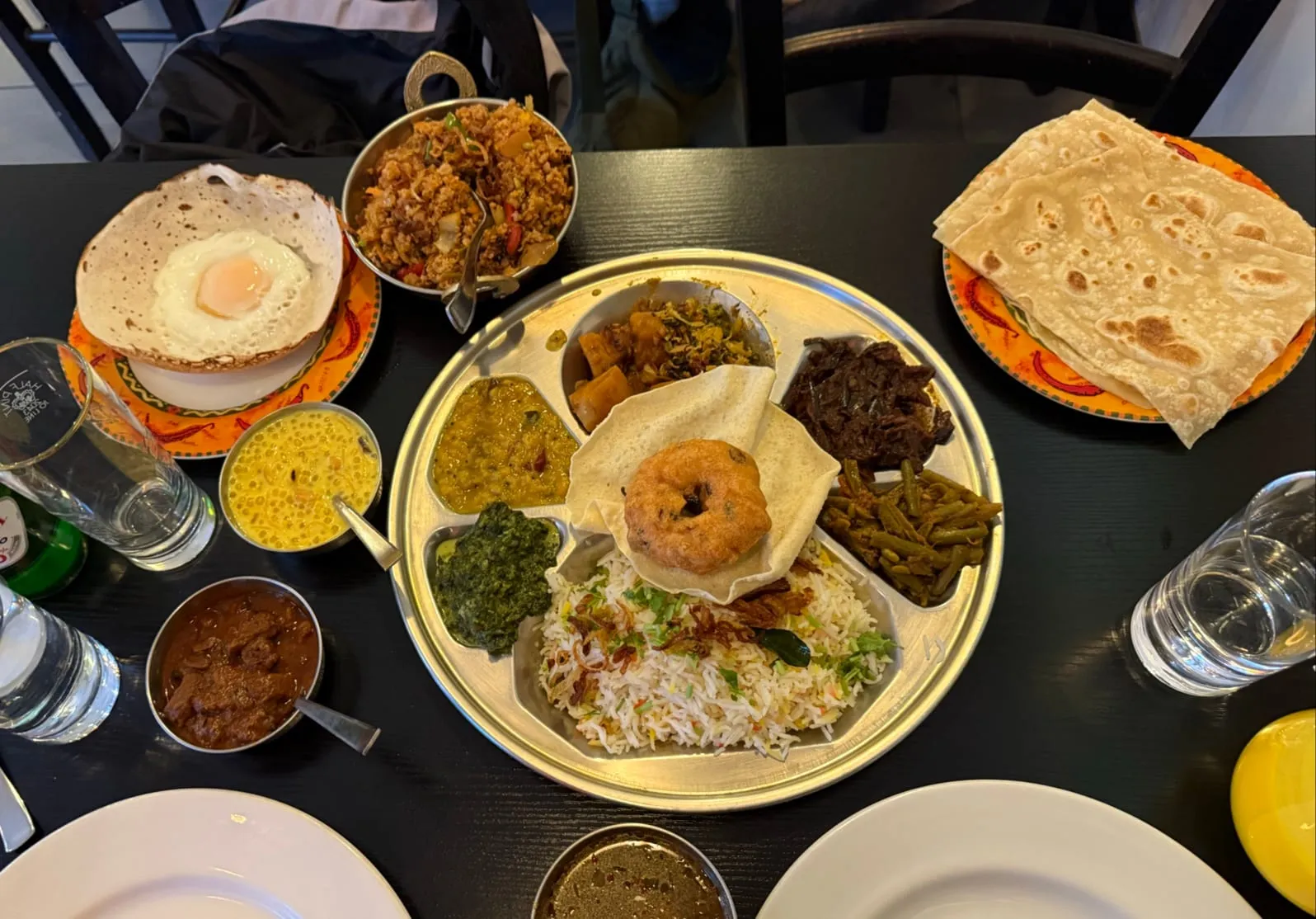 A table of dishes: an egg hopper, a bowl-shaped pancake with a lacy rim and a fried egg set into the base; a steel plate of rice with dal, a dark green curry, green beans, a diced vegetable curry and a ring-shaped lentil vadai; a steel bowl of kottu; and a stack of flatbreads on chilli-print plates