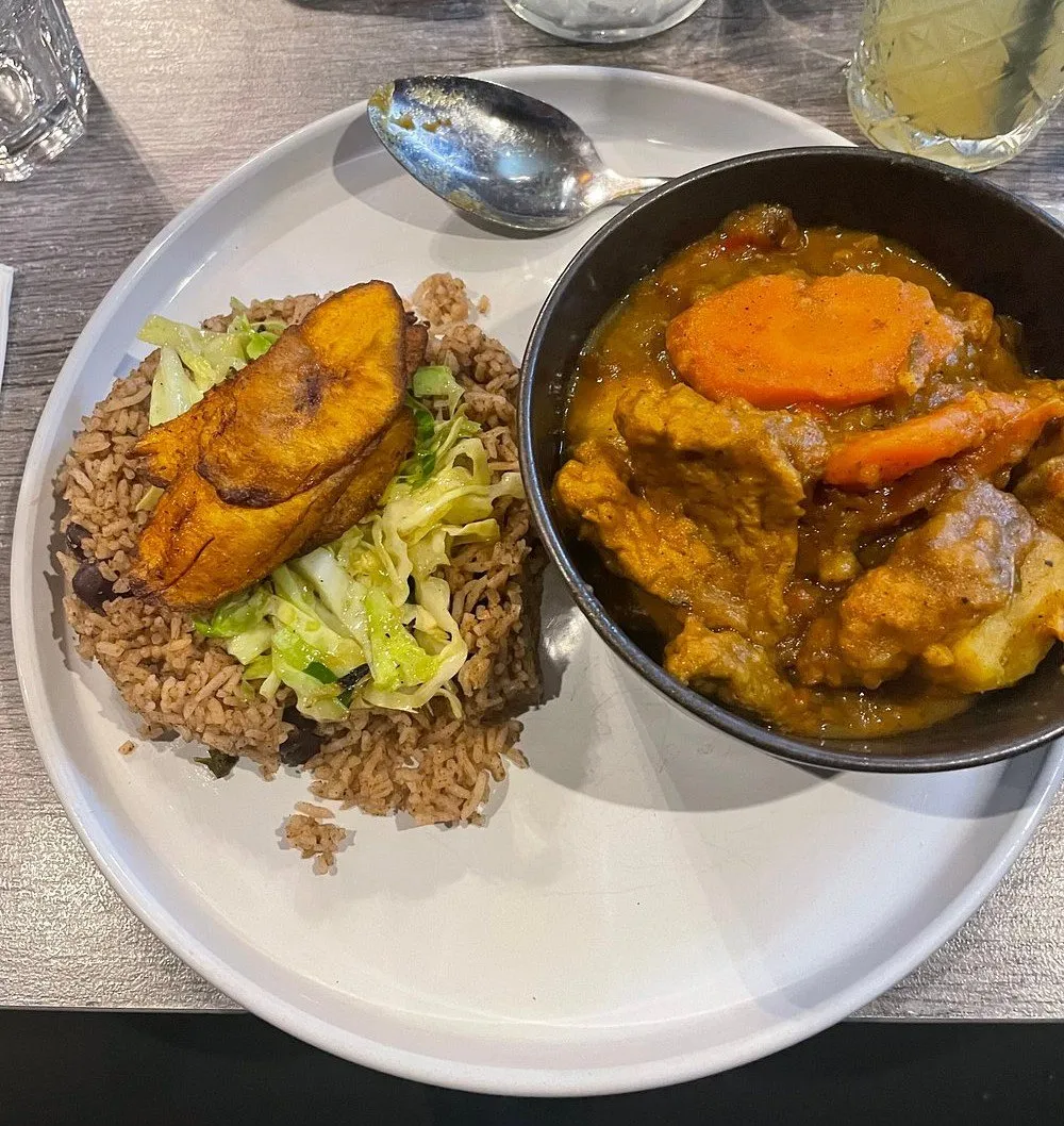 A plate of vegan Caribbean food - a curry with carrot and dumplings in a black bowl, beside rice and peas topped with fried plantain and steamed cabbage
