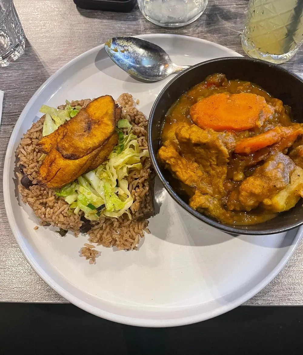 A curry with carrot in a black bowl beside rice and peas topped with a wedge of fried plantain and steamed cabbage, on a white plate