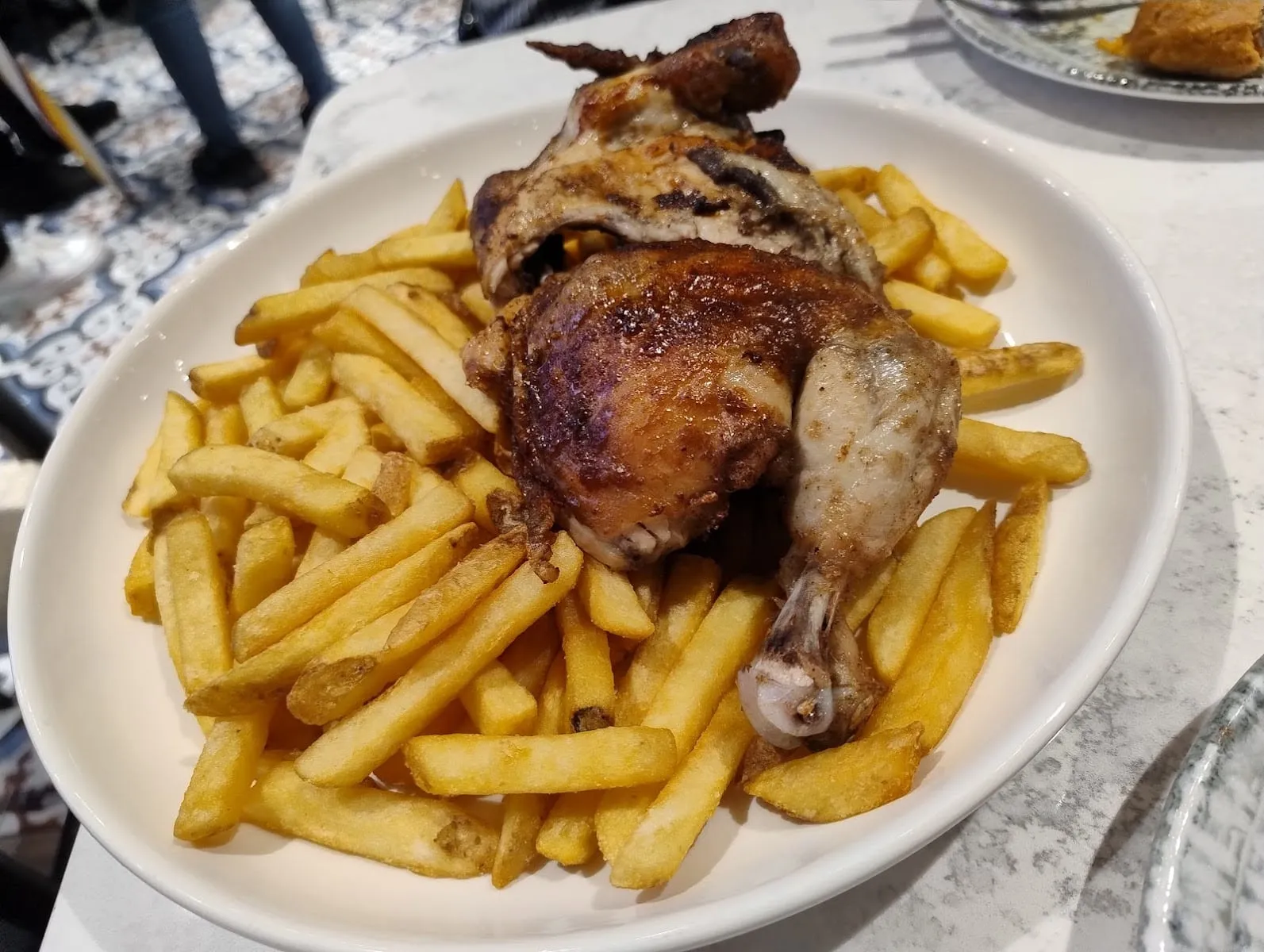 Half a charcoal-roasted chicken, the skin browned and blistered, sitting on a bed of chips on a white oval plate