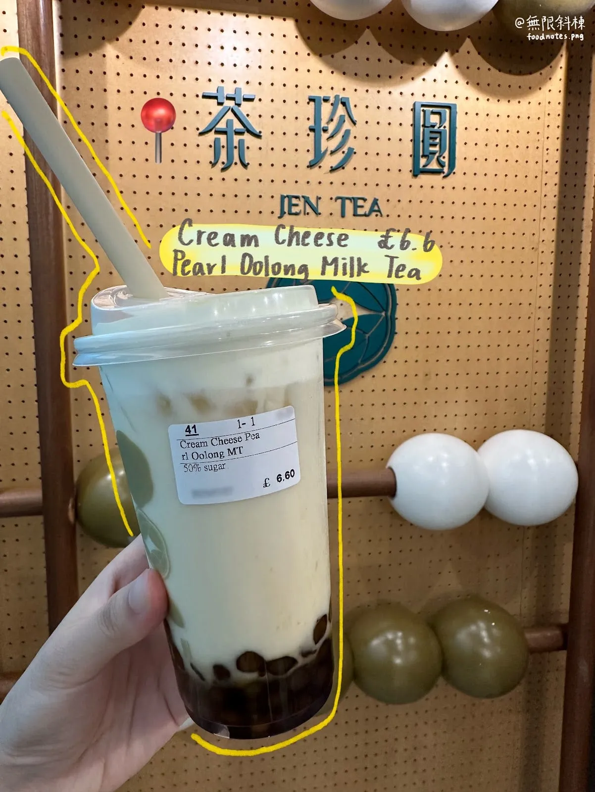 A cream cheese pearl oolong milk tea held up in front of Jen Tea's pegboard wall, with the price, £6.60, written on the board behind