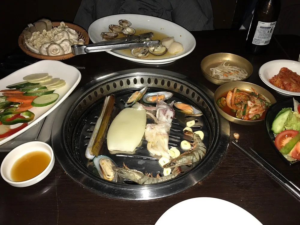 Seafood grilling on a sunken table grill at Jin Go Gae - squid, a razor clam, mussels and a whole prawn with sliced garlic, ringed by banchan, pickled vegetables and a bowl of kimchi