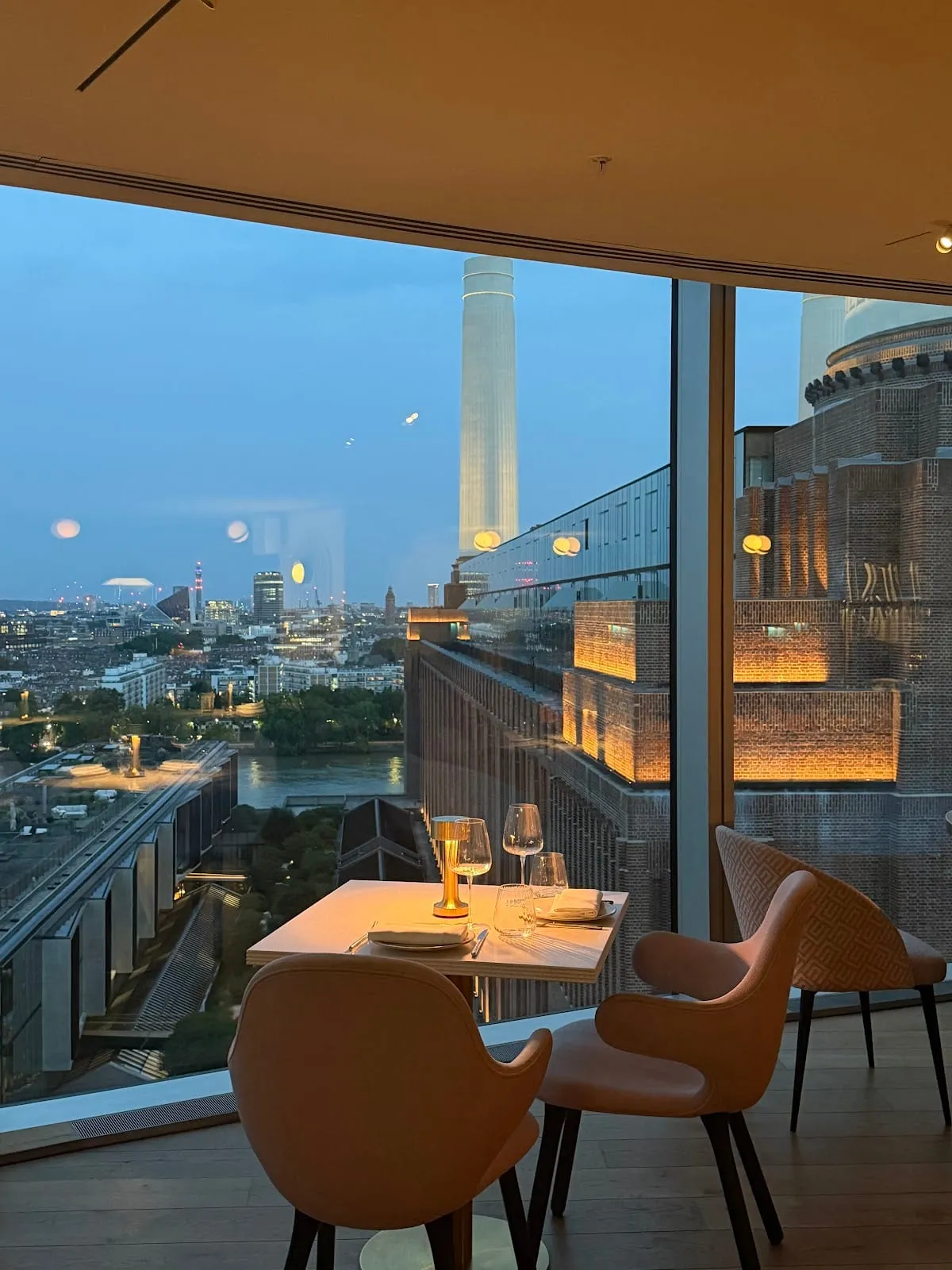 A table for two at Joia beside a floor-to-ceiling window, with a chimney of Battersea Power Station and the London skyline outside at dusk