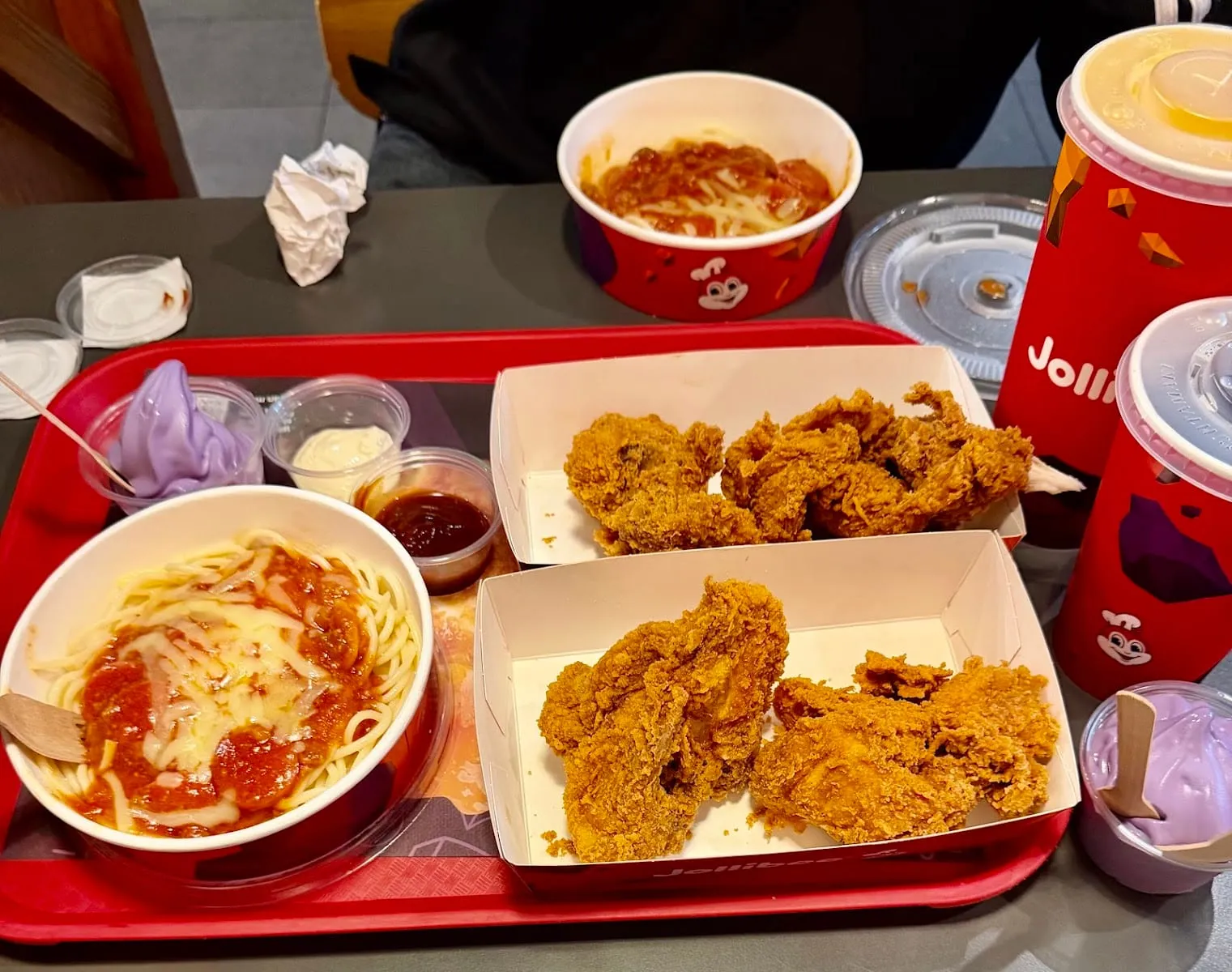 A red Jollibee tray holding two boxes of fried chicken, a bowl of spaghetti under red sauce and melted cheese, pots of gravy and mayonnaise, two cups of purple soft serve and branded drinks cups