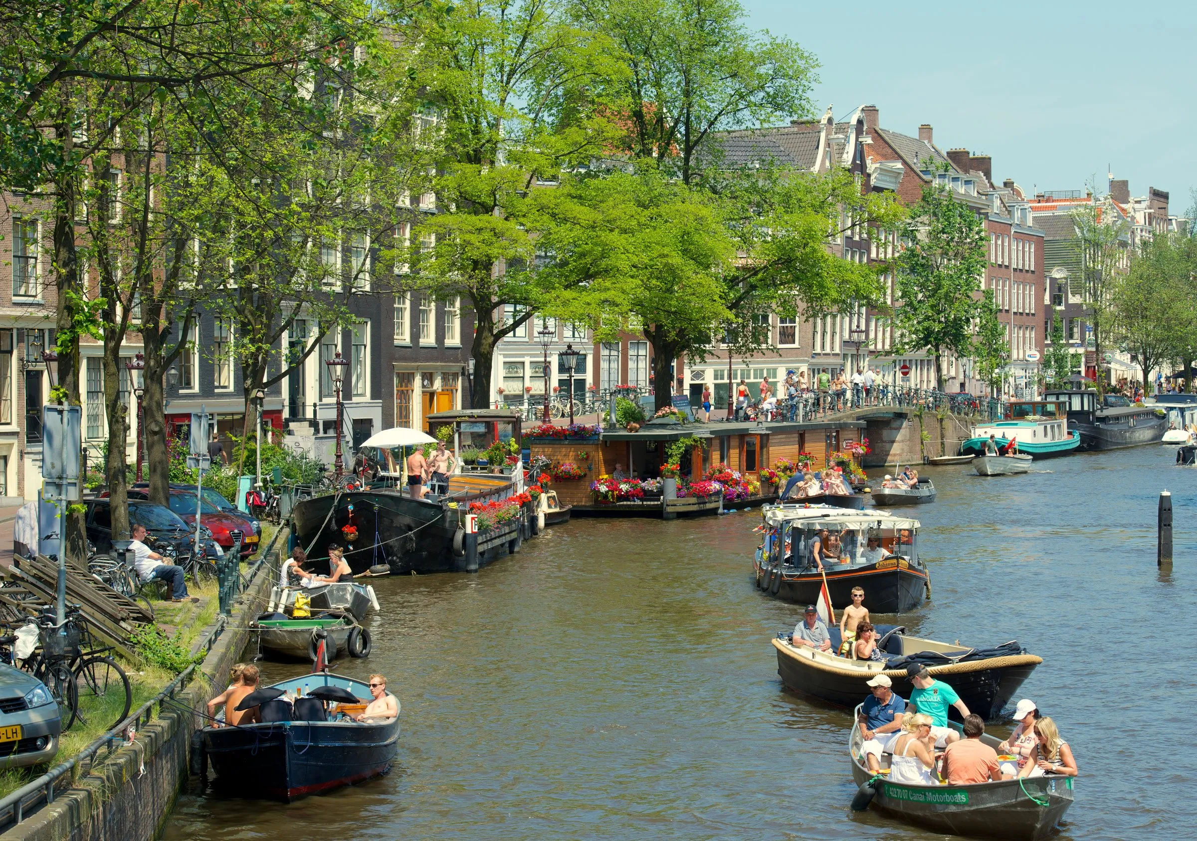 Small motorboats and houseboats moored along a tree-lined Jordaan canal in Amsterdam, with gabled houses and bicycles along the towpath