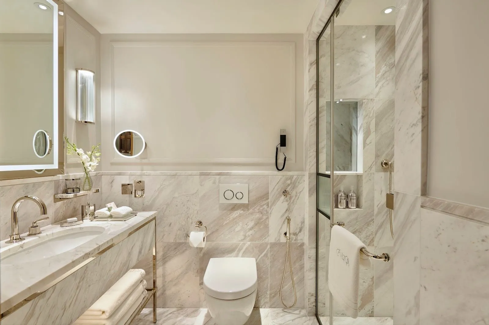A guest room bathroom at Jumeirah Carlton Tower in grey-veined marble, with a walk-in shower behind a glass door, a wall-hung toilet and a basin on a marble washstand