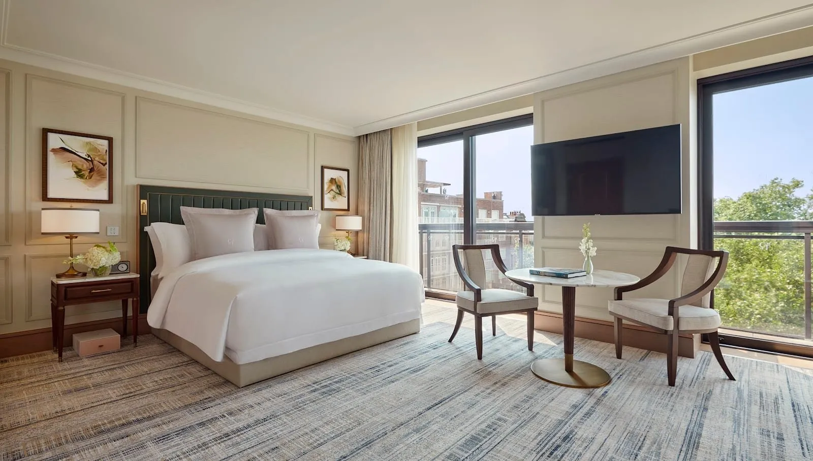 A Junior Suite at Jumeirah Carlton Tower: a king bed with a dark green leather headboard, cream panelled walls, a round marble table with two chairs, and floor-to-ceiling windows onto a balcony and treetops