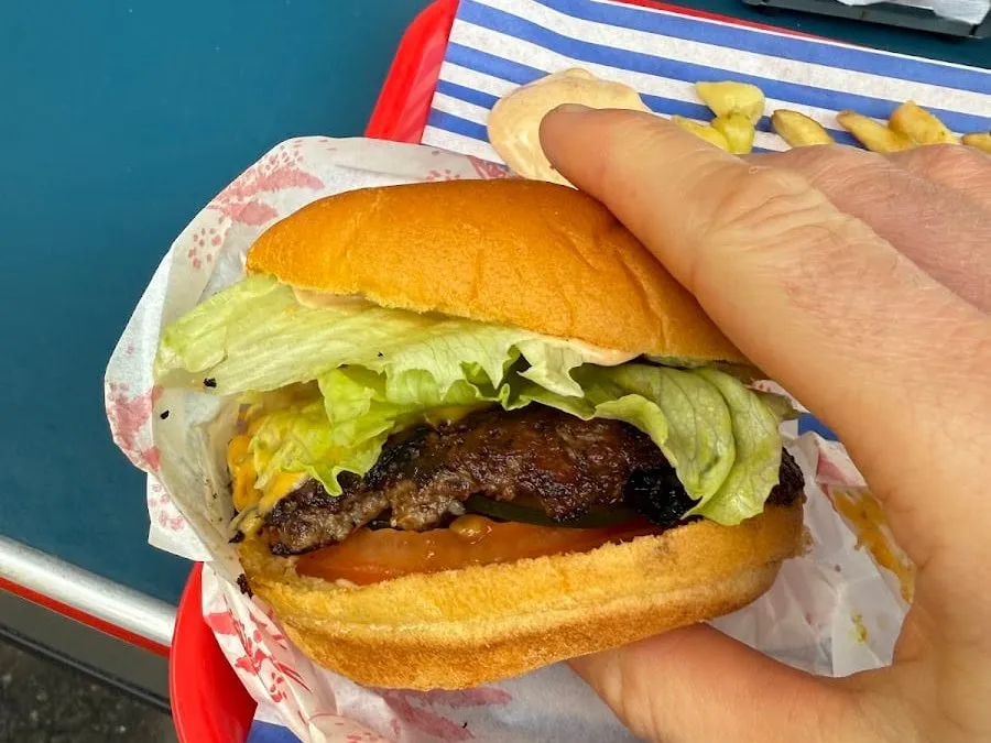 A Jupiter Burger cheeseburger held in one hand over a red tray lined with blue-and-white striped paper: a charred beef patty with melted cheese, iceberg lettuce, tomato, a pickle slice and pink sauce in a soft yellow potato roll