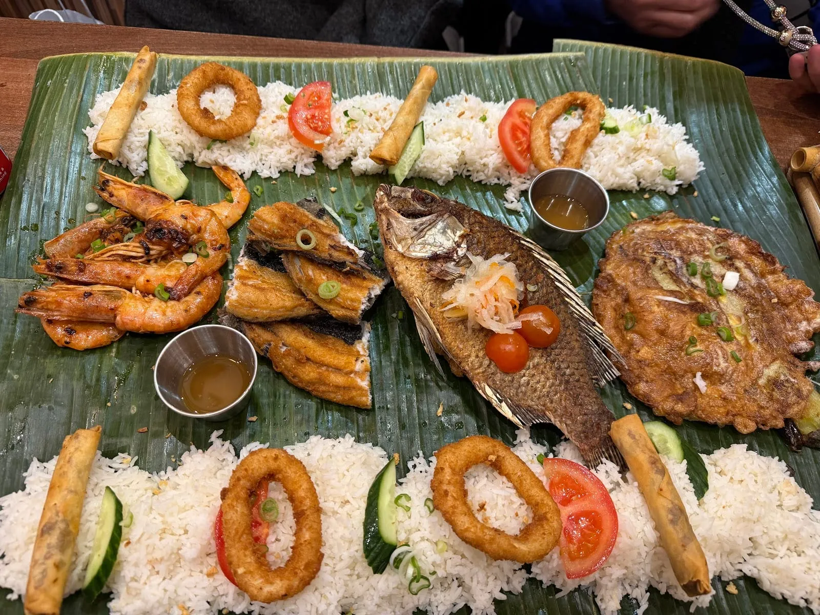 A banana leaf running down the middle of a table, laid with mounds of rice, spring rolls, onion rings, tomato and cucumber slices, grilled prawns, split fried fish, a whole fried fish under pickled papaya and a flat golden omelette, with diners' hands at the edge of the frame