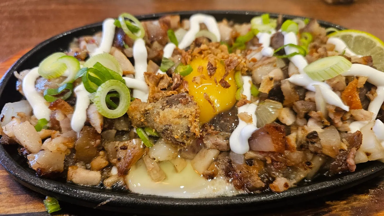 Sisig served on a black cast-iron sizzling plate: chopped crisp pork with spring onions, a drizzle of mayonnaise, a raw egg yolk set in the centre and a wedge of lime