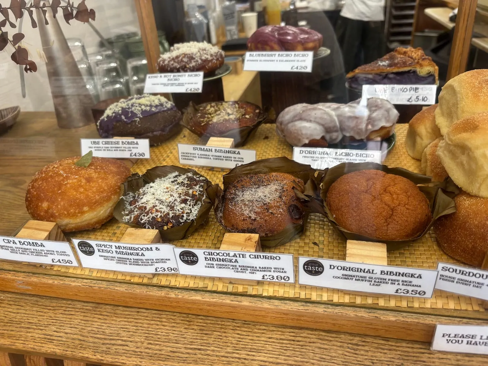 A glass bakery counter of Filipino bakes on woven mats, each behind a printed label: bibingka in banana leaves, glazed bicho-bicho doughnuts, an ube cheese bomba, a slice of purple-filled pie and a stack of pandesal