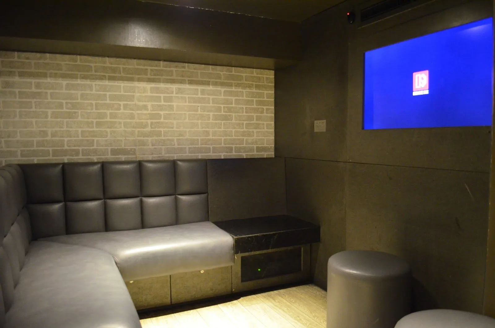 A small karaoke room at Karaoke Box Soho, with a grey padded corner sofa, a stool, a brick-effect wall and a screen showing the Karaoke Box logo