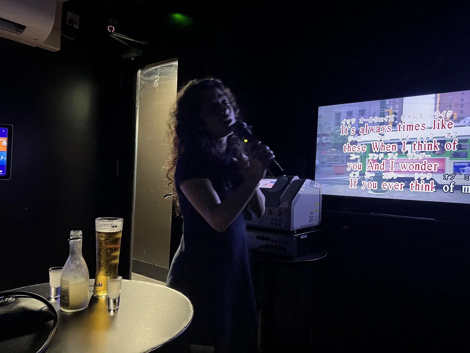 A woman singing in a dark karaoke room at Karaoke Epoc, with lyrics in English and Japanese on the screen and drinks on a small table