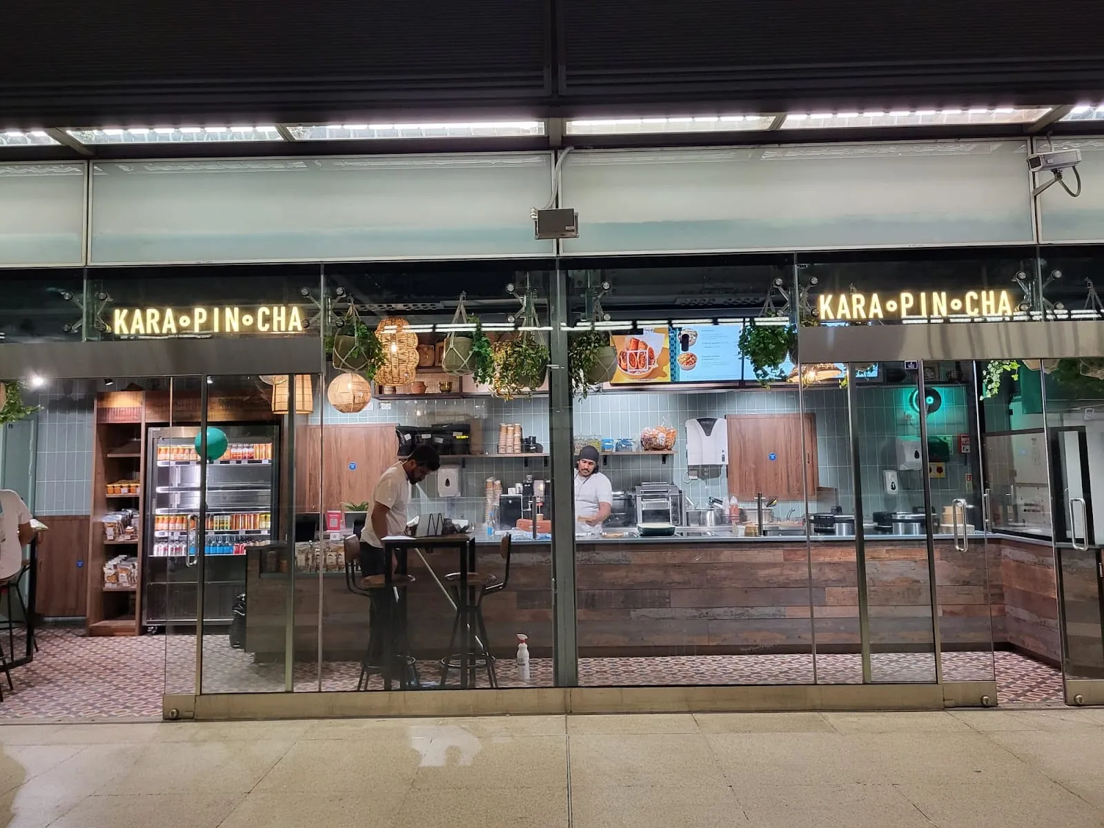A glass-fronted counter unit with the Karapincha name lit in yellow letters above it, two cooks working an open kitchen behind a reclaimed-wood counter, pale green tiling, hanging plants and rattan pendant lamps