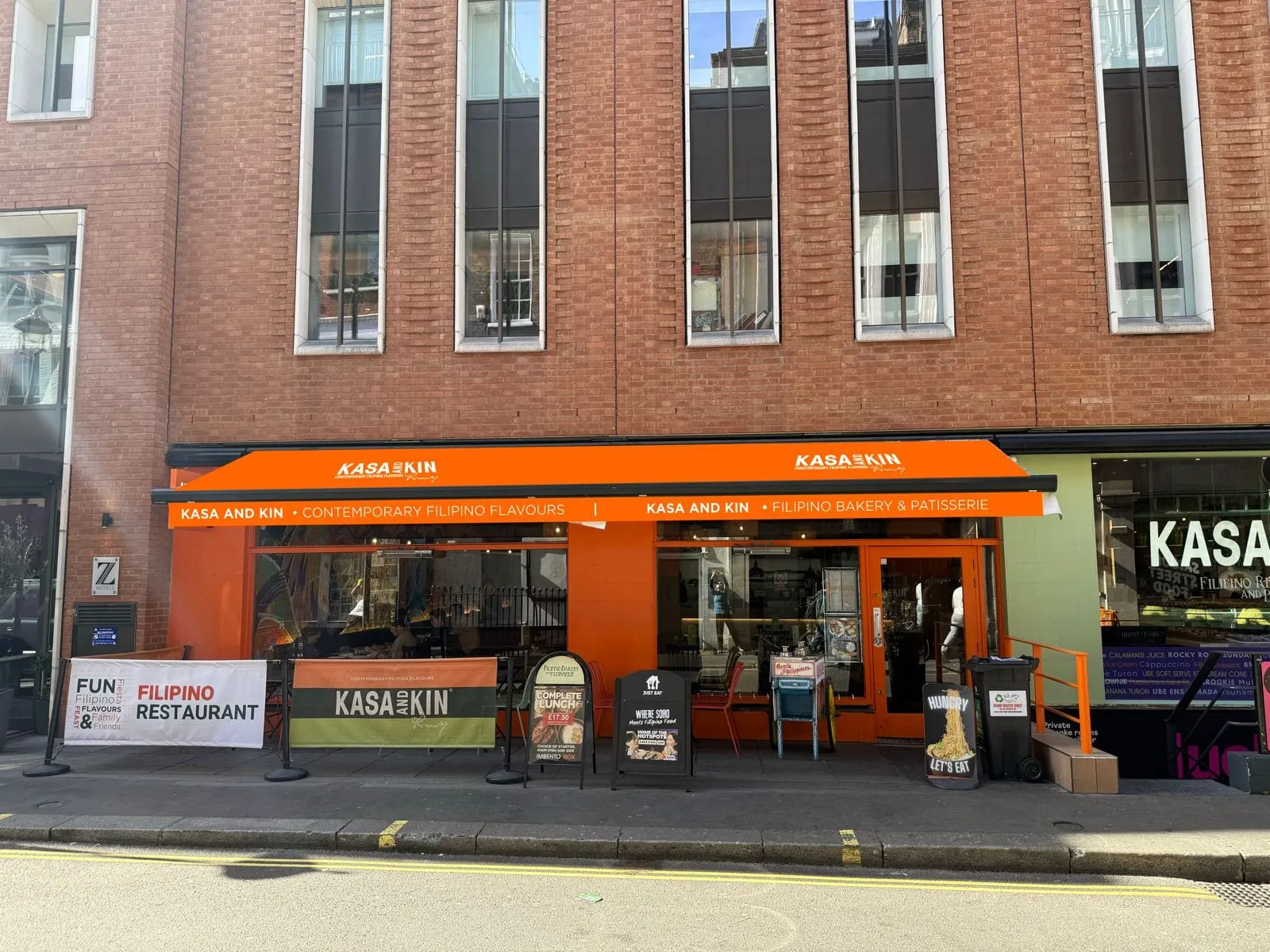 The Kasa and Kin shopfront under a bright orange awning reading "contemporary Filipino flavours" on one side and "Filipino bakery and patisserie" on the other, with pavement barriers, A-boards and a menu case beside the door