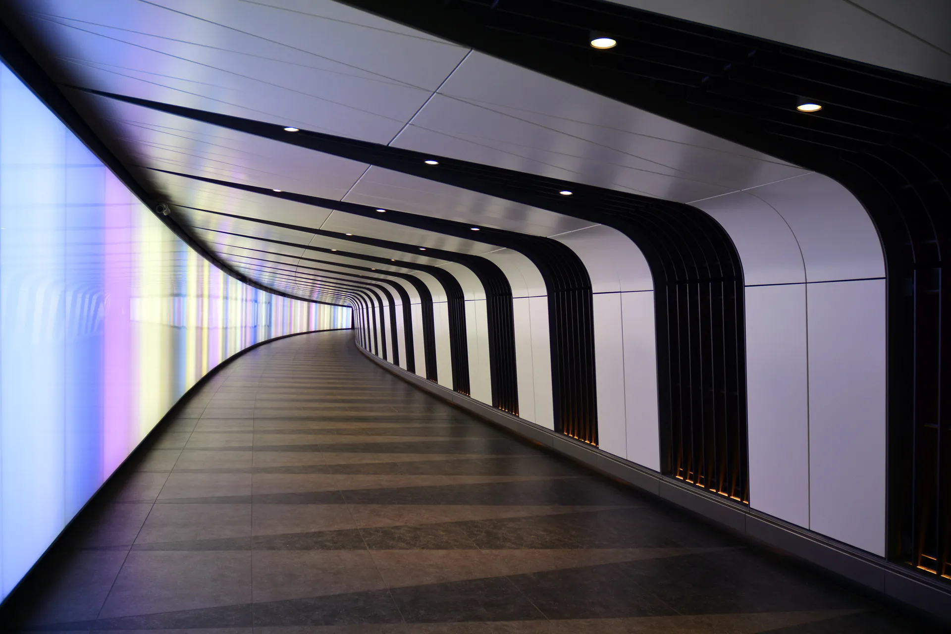 The curved wall of the King's Cross Light Tunnel glowing blue, lilac and yellow, opposite a wall of black and white vertical bands