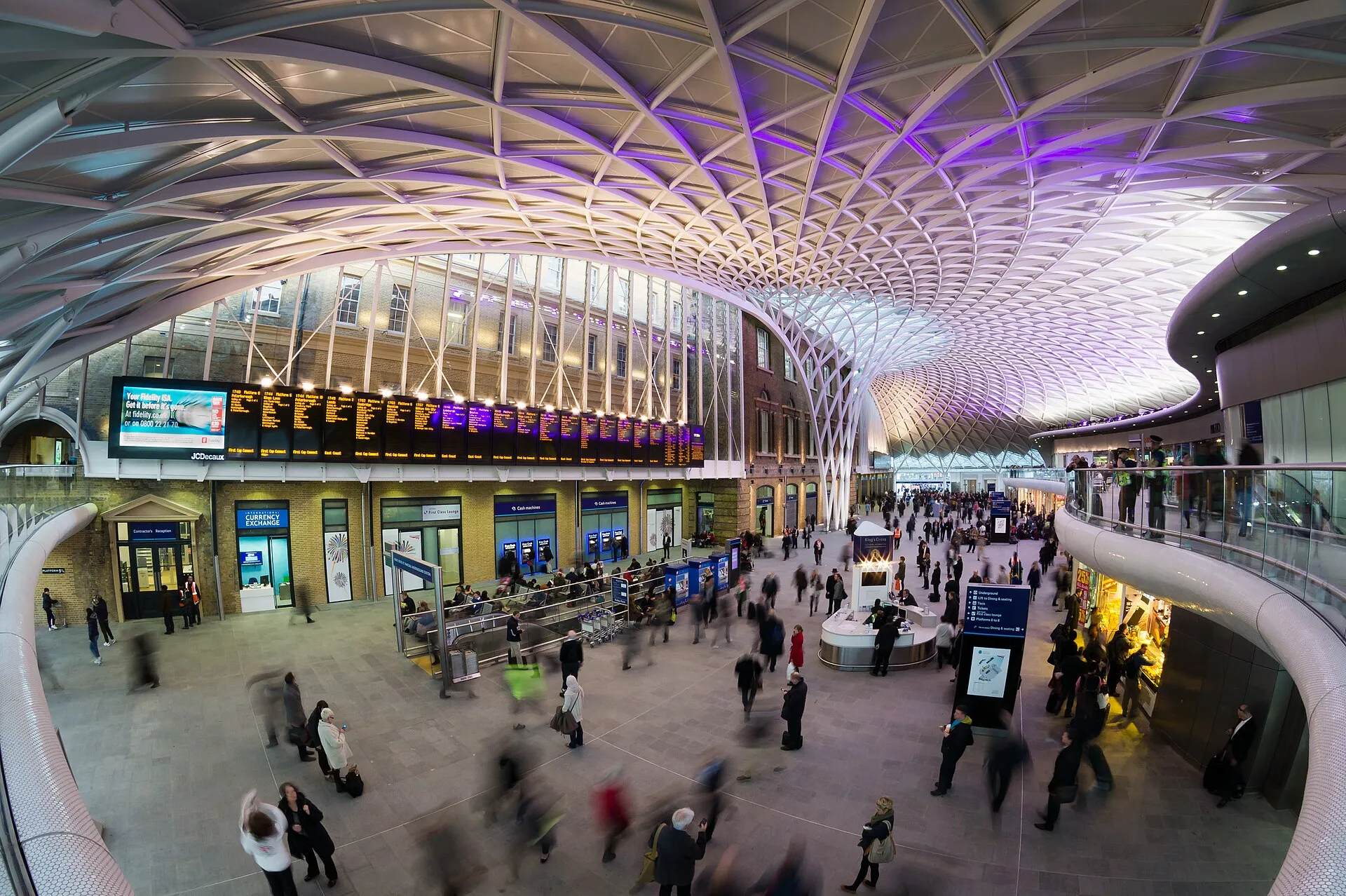 The western concourse at King's Cross under its fanned white lattice roof, departure boards along the left-hand wall and travellers crossing the floor with cases
