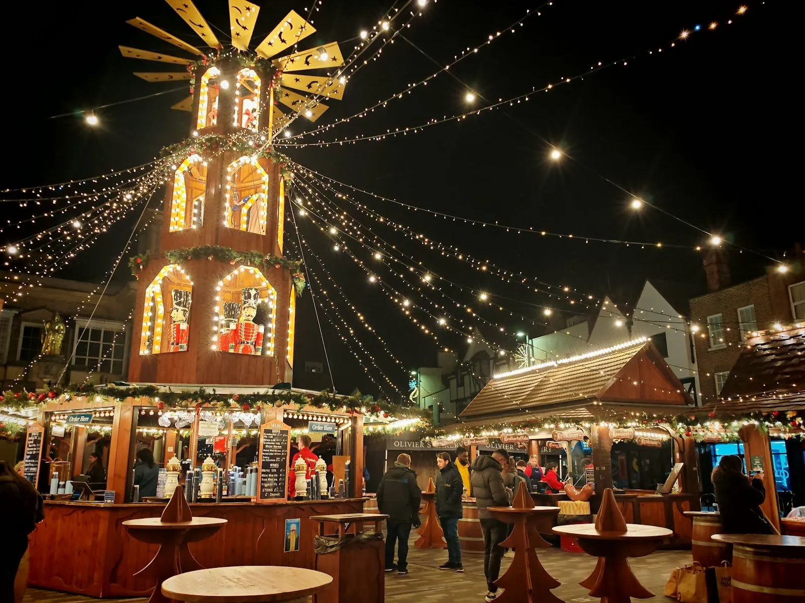 A tall wooden Christmas pyramid with nutcracker soldiers and a star-cut sail above a bar at night in Kingston's Ancient Market Place, strings of lights running from its top to the chalets, and the gilded statue on the Market House at left