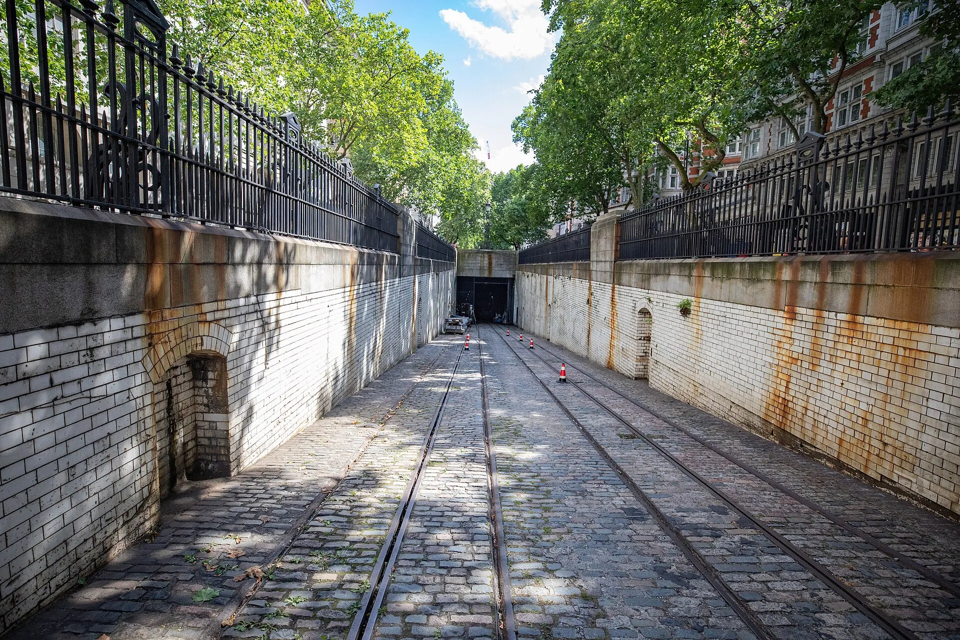 A cobbled ramp with tram rails running down between white-tiled walls topped with iron railings, towards a dark tunnel mouth
