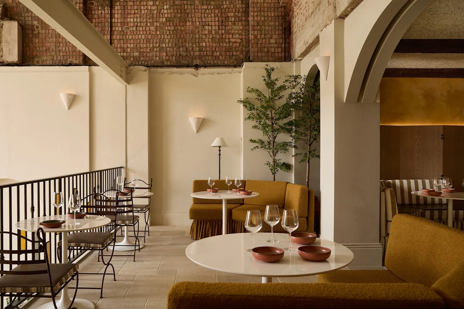 A high-ceilinged dining room with exposed brick above, round tables and mustard banquettes