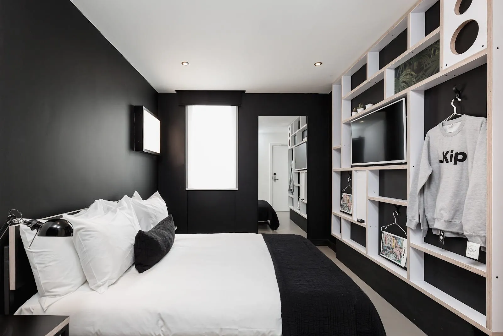 A double room at Kip Hotel with a black wall, white bedding and open shelving holding a TV and a Kip sweatshirt