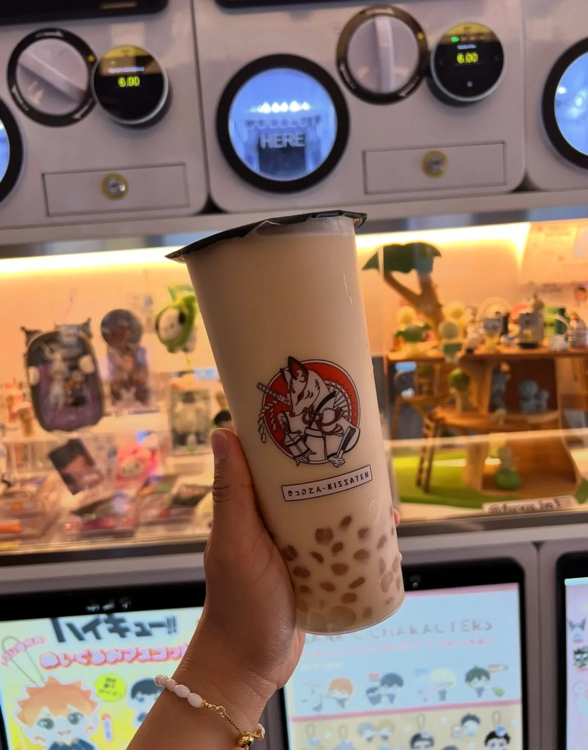 A cup of milk tea with amber pearls held up in front of Kissaten's glowing wall of gachapon capsule machines