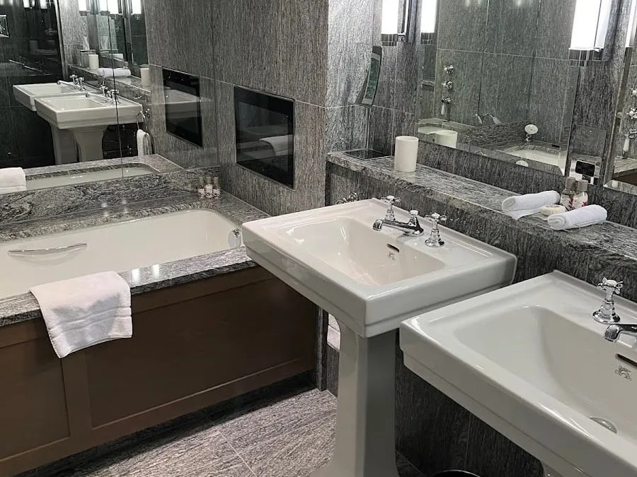A bathroom at the Knightsbridge Hotel in grey granite, with a bath panelled in dark oak, two white pedestal basins and a television set into the wall