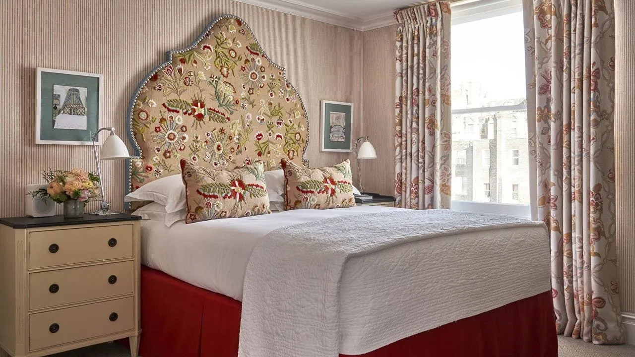 A bedroom at the Knightsbridge Hotel with a tall arched headboard embroidered with red and green flowers, matching cushions, a red valance, pinstriped walls, floral curtains and a sash window