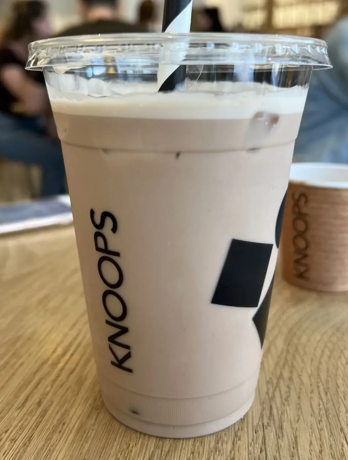 A Knoops iced chocolate in a branded clear cup with a striped paper straw, on a light wood table