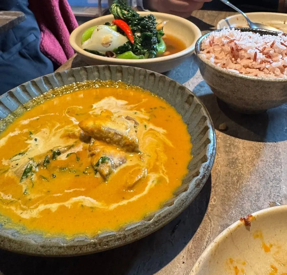 A rich orange coconut curry in a fluted stoneware bowl, beside a bowl of red rice and a dish of greens with whole red chilli