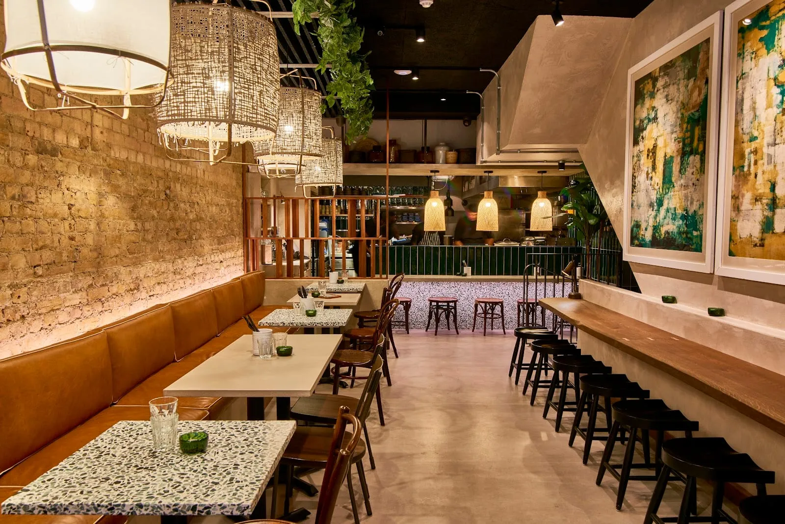 A narrow dining room: a tan leather banquette down an exposed brick wall, terrazzo table tops with bentwood chairs, woven rattan pendant shades, and on the other side a long wooden counter of black stools facing an open kitchen behind green tiling