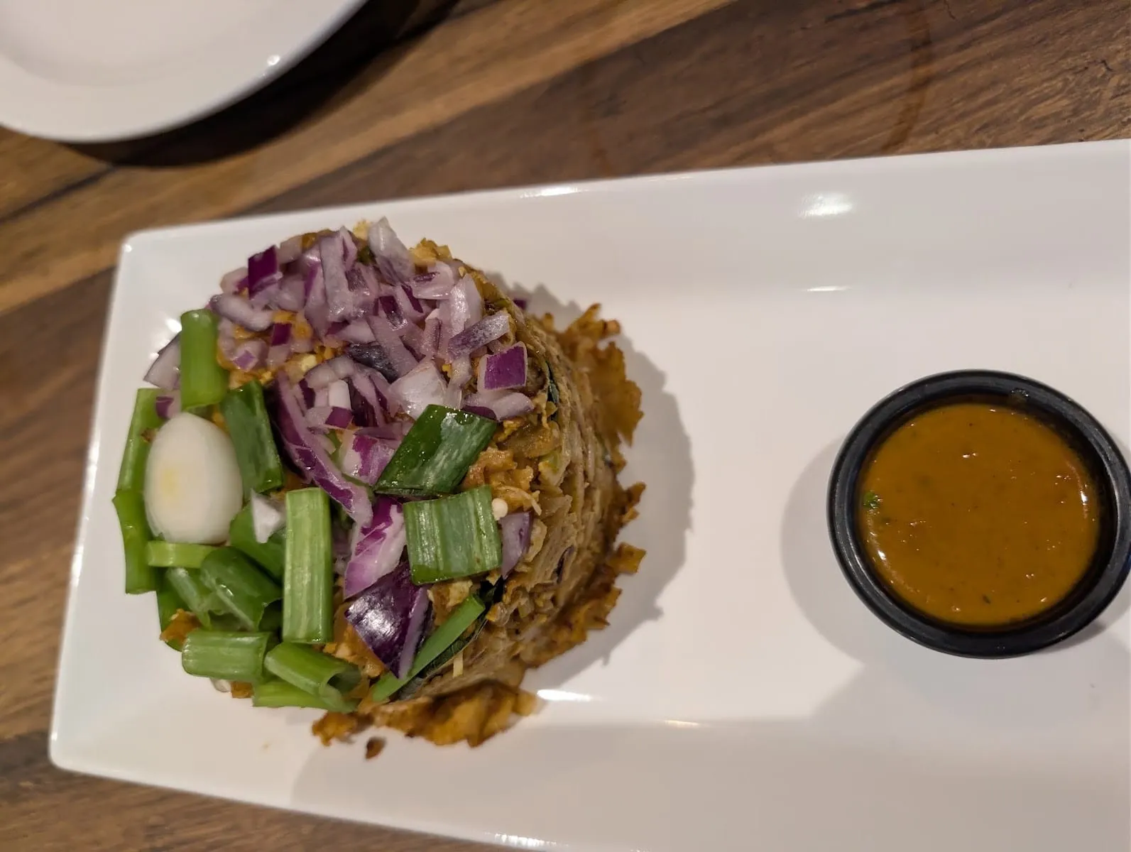 A domed mound of kothu on a long white plate — roti shredded fine on the griddle, scattered with chopped red onion and cut spring onion and topped with a slice of boiled egg — beside a small black pot of thin curry sauce