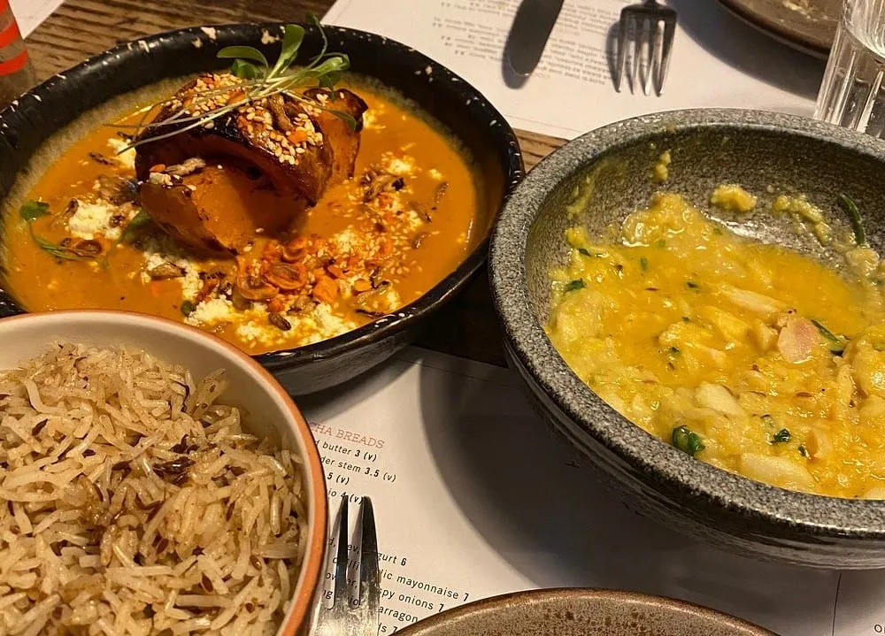 Two small plates at Kricket — roast pumpkin in a makhani sauce with crumbled cheese and pumpkin seeds, and a dal — beside a bowl of cumin rice, with the printed menu visible underneath