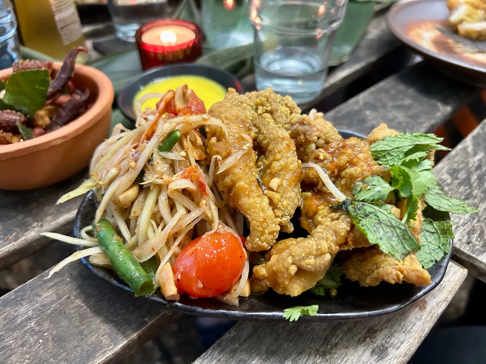 Fried chicken pieces beside a green papaya salad with cherry tomatoes, on a dark plate