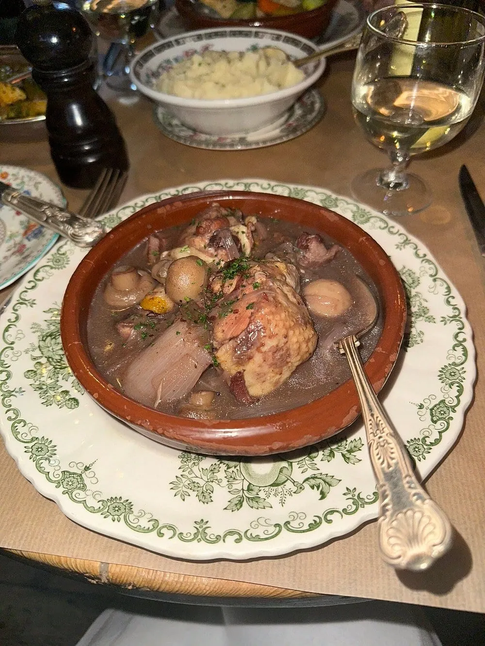 Coq au vin in a terracotta dish set on a green-patterned plate, with mashed potato in a floral bowl, a glass of white wine and a wooden pepper mill on the table