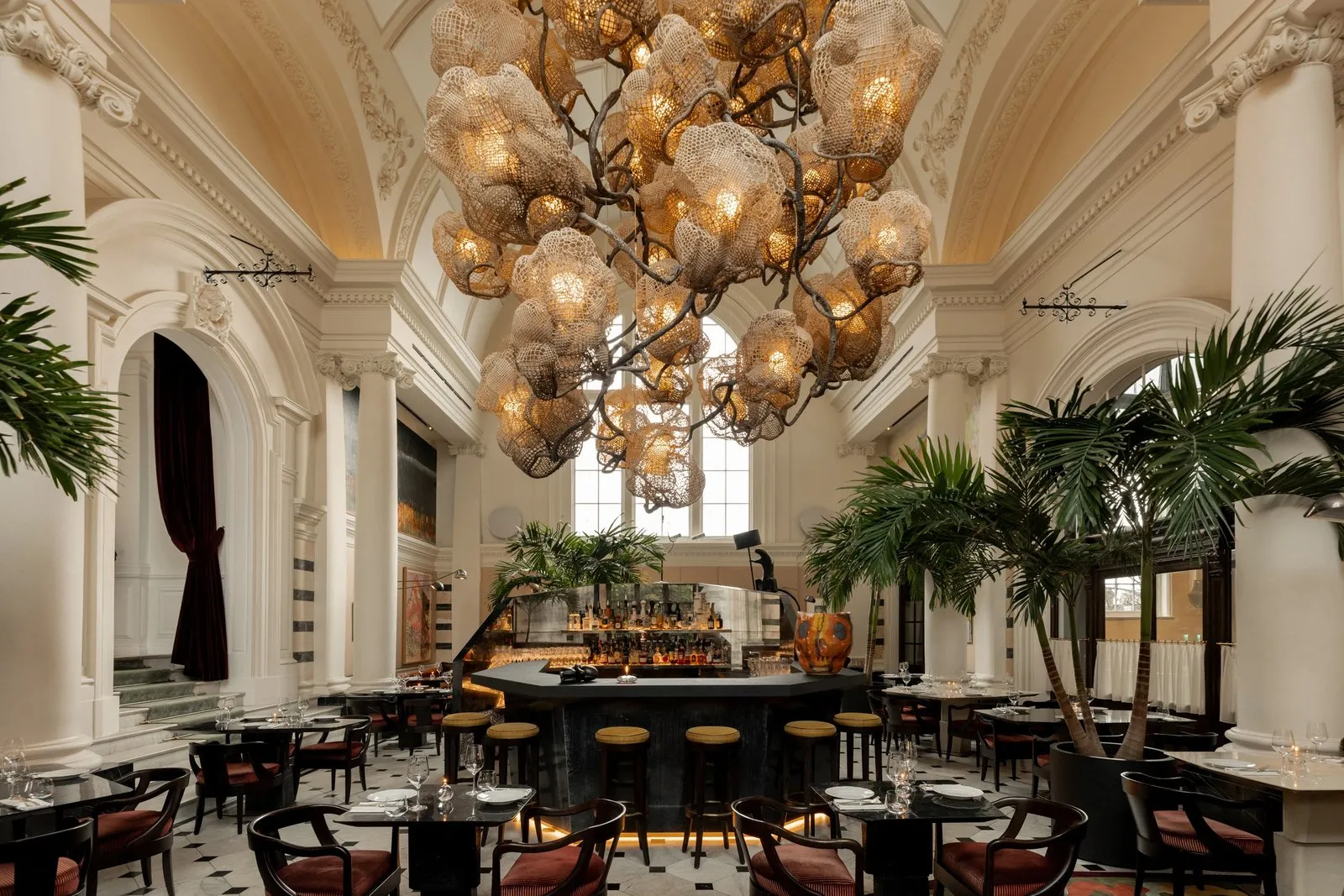 A vaulted dining room at Ladbroke Hall with a large sculptural chandelier, palm trees, a central bar and tables laid for dinner