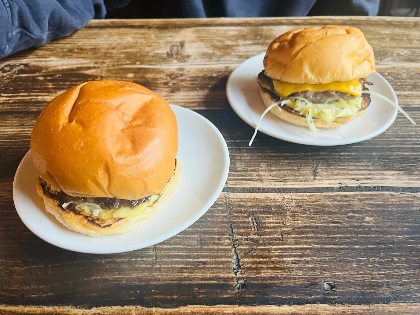 Two Lagom burgers on white plates on a worn wooden pub table, the nearer one closed, the further one showing a beef patty, a slice of melted cheese and shredded slaw