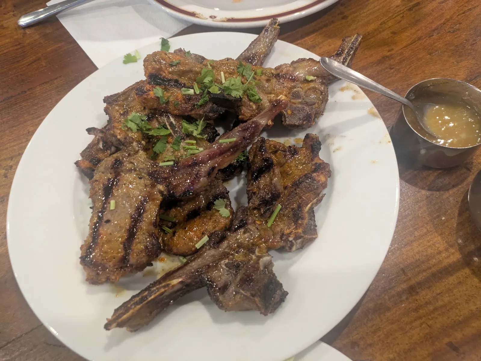 Charred lamb chops on a plate at Original Lahore Kebab House, with a metal pot of dip alongside