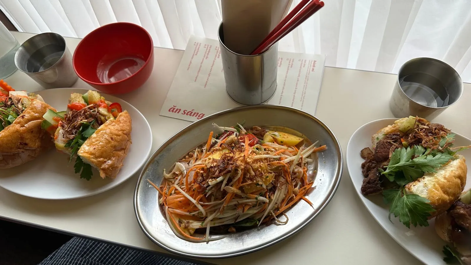 A banh mi sandwich and a green papaya-style salad laid out on a table at Lai Rai, with steel cups