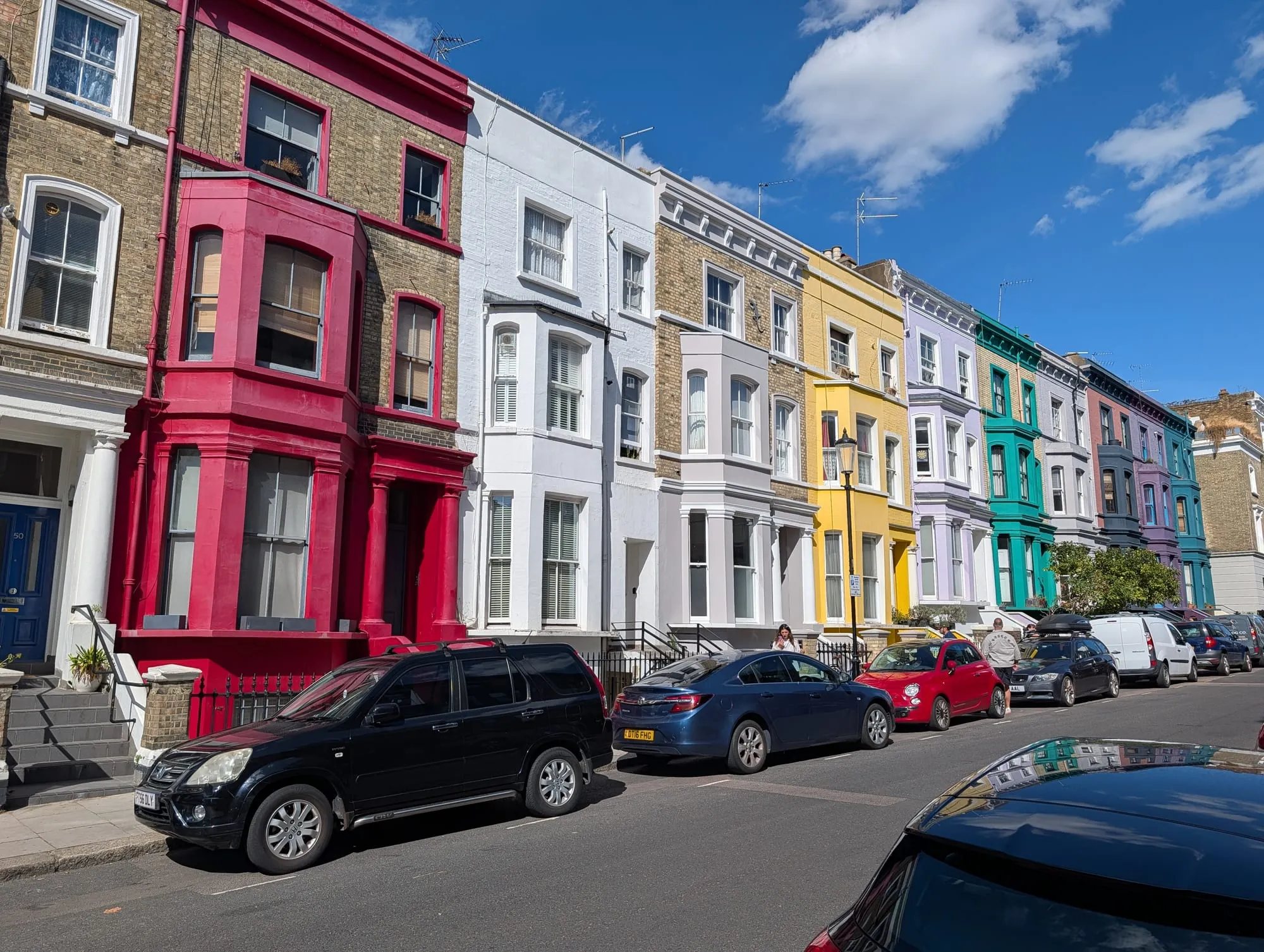 A terrace on Lancaster Road painted deep pink, white, blue and green, with cars parked along the kerb