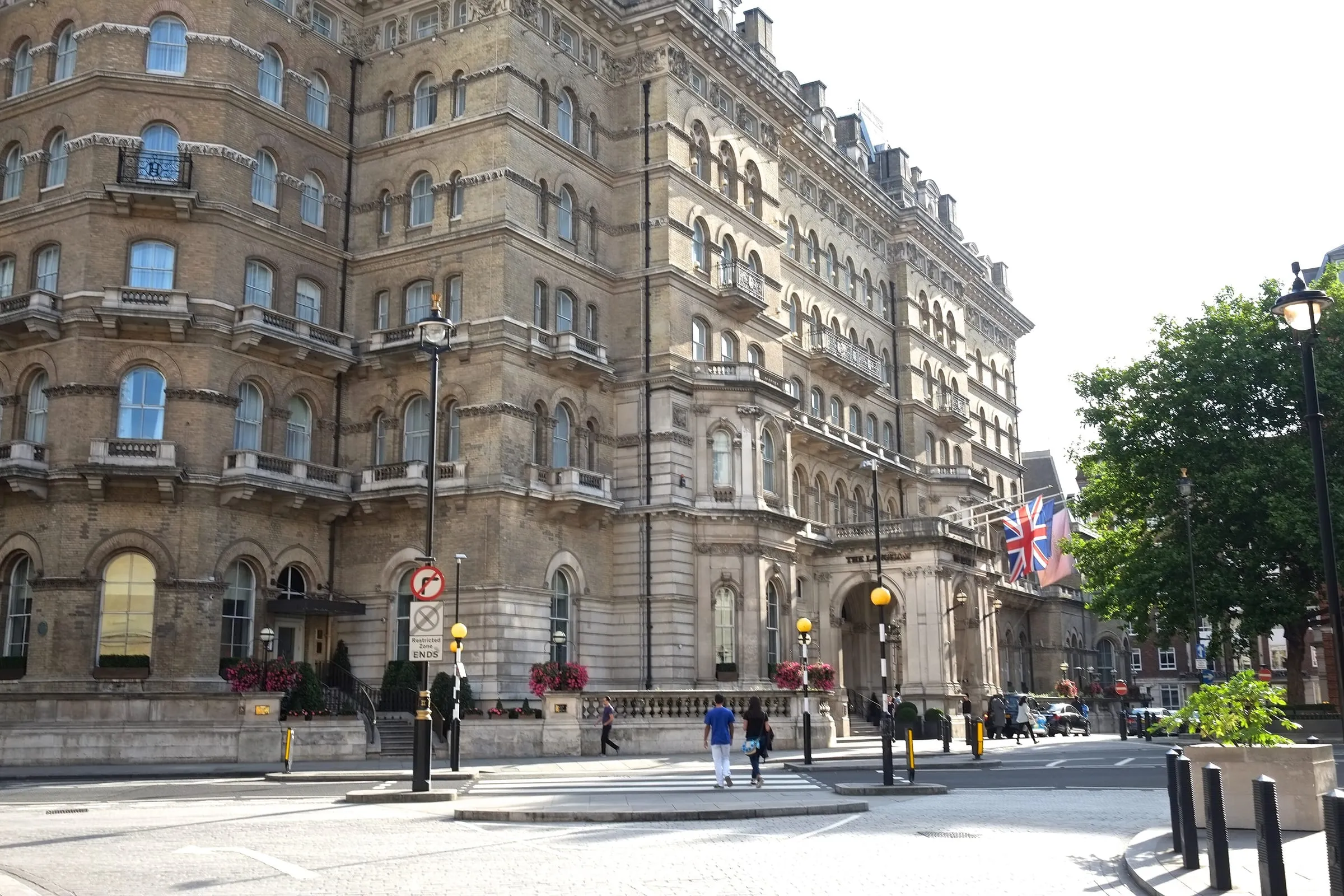 The Langham's Victorian stone frontage on Portland Place, with balconied upper floors, Union flags over the arched entrance and pedestrians crossing the road in front