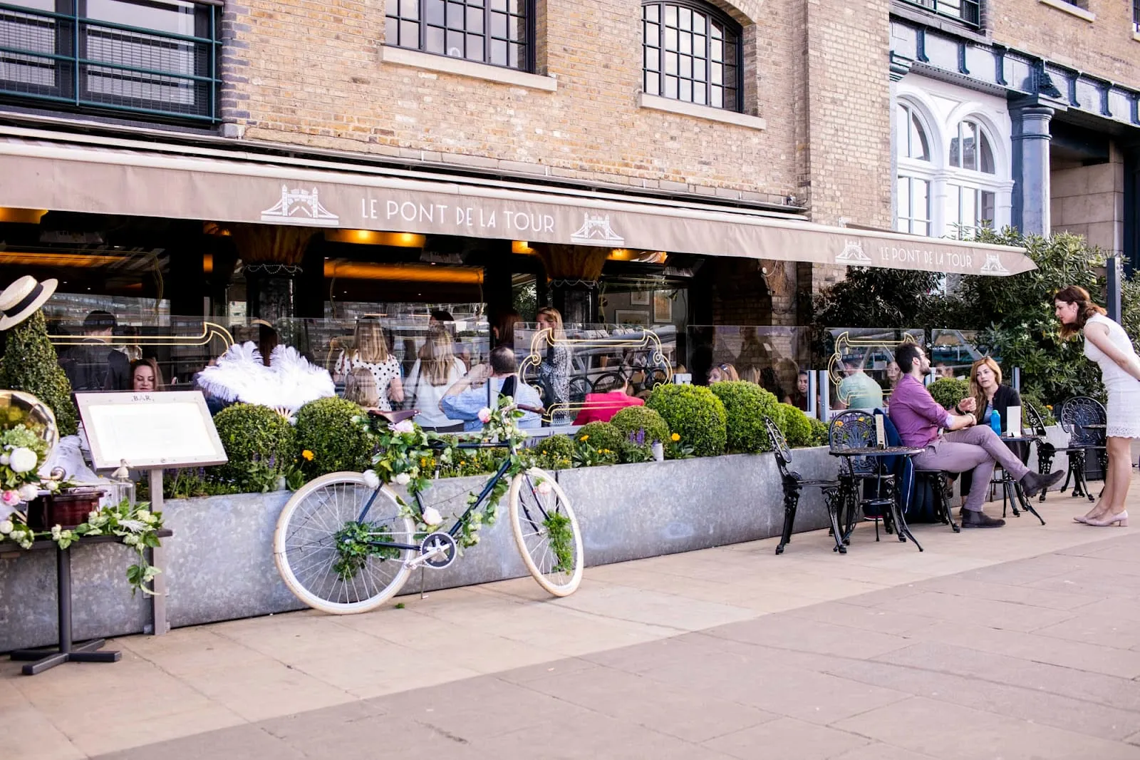 The terrace at Le Pont de la Tour on Butlers Wharf: an awning carrying the restaurant's name and a Tower Bridge motif, cast-iron café tables on the promenade, clipped box in long zinc planters, and a flower-dressed white bicycle propped against them