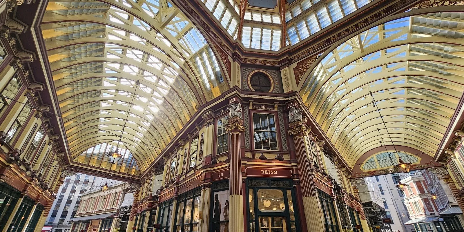 Looking up at the crossing in the middle of Leadenhall Market: cream and maroon ironwork, glass roofs meeting under a dome, and silver dragons on the columns either side of the Reiss shop at number 26