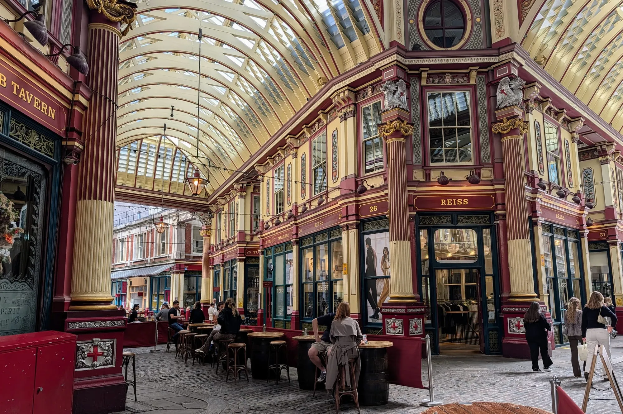 The painted maroon, cream and gold arcade of Leadenhall Market under its glass roof, with the Lamb Tavern on the left and drinkers at barrel tables along the lane