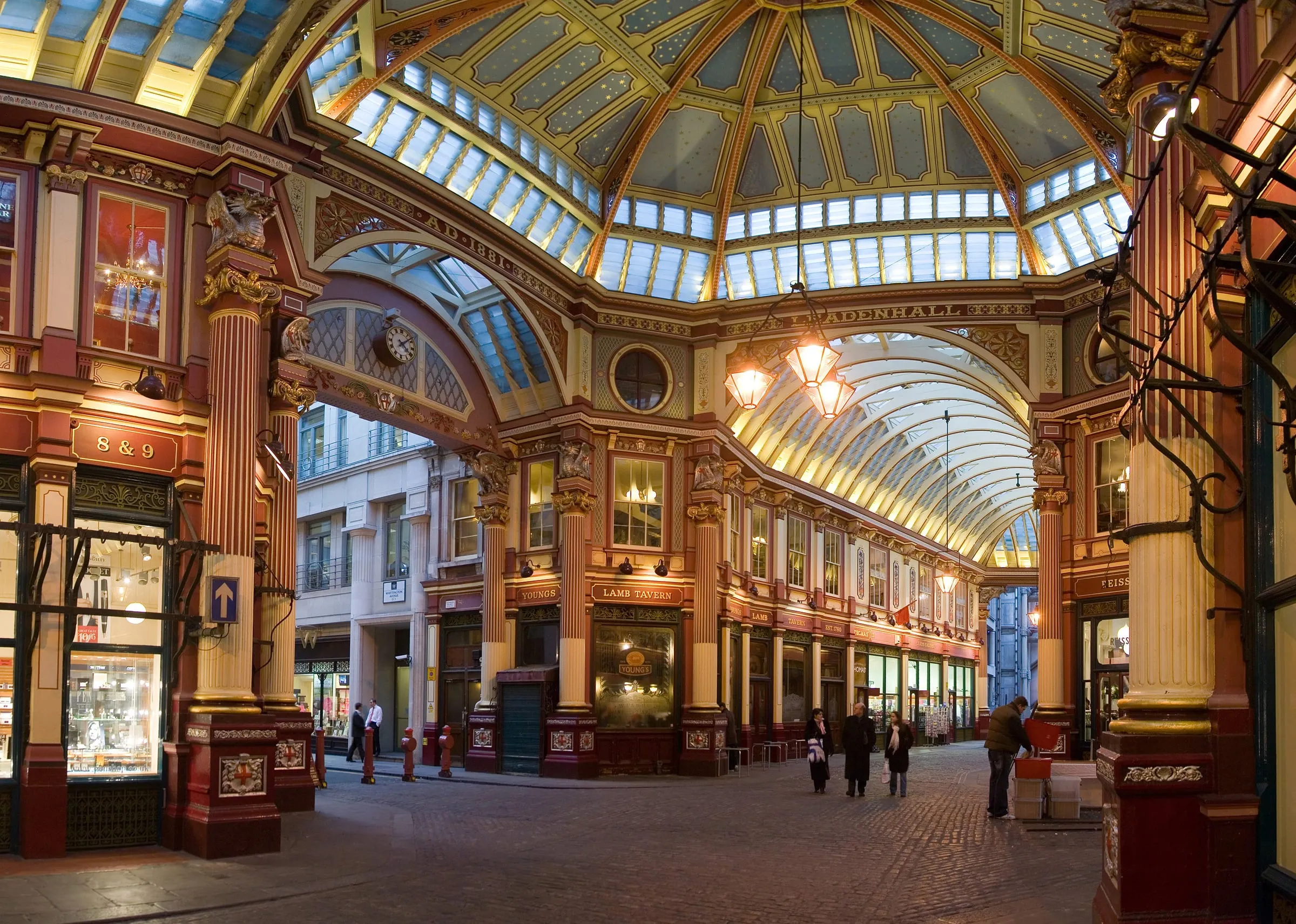 The crossing at the centre of Leadenhall Market, its maroon and cream columns, gilded dome and glass roofs lit up, with the Lamb Tavern at the end of the cobbled lane