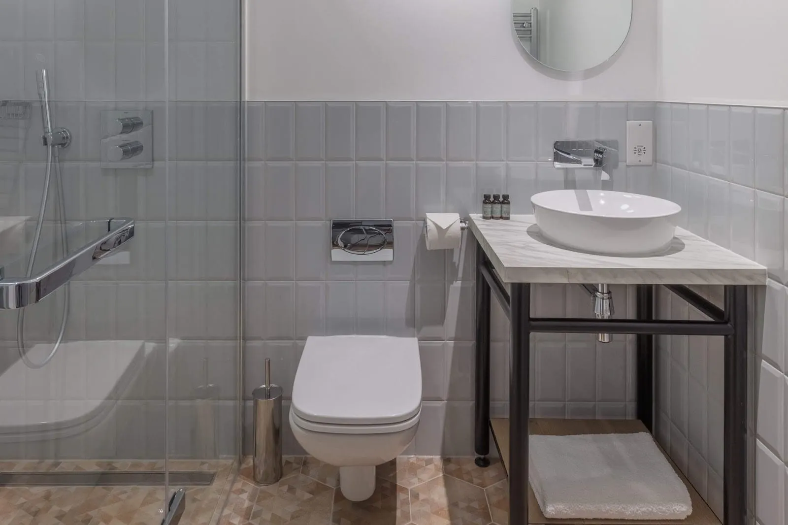A bathroom at Leman Locke with grey bevelled tiles, a walk-in shower behind a glass screen, a wall-hung toilet and a bowl basin on a marble-topped black stand