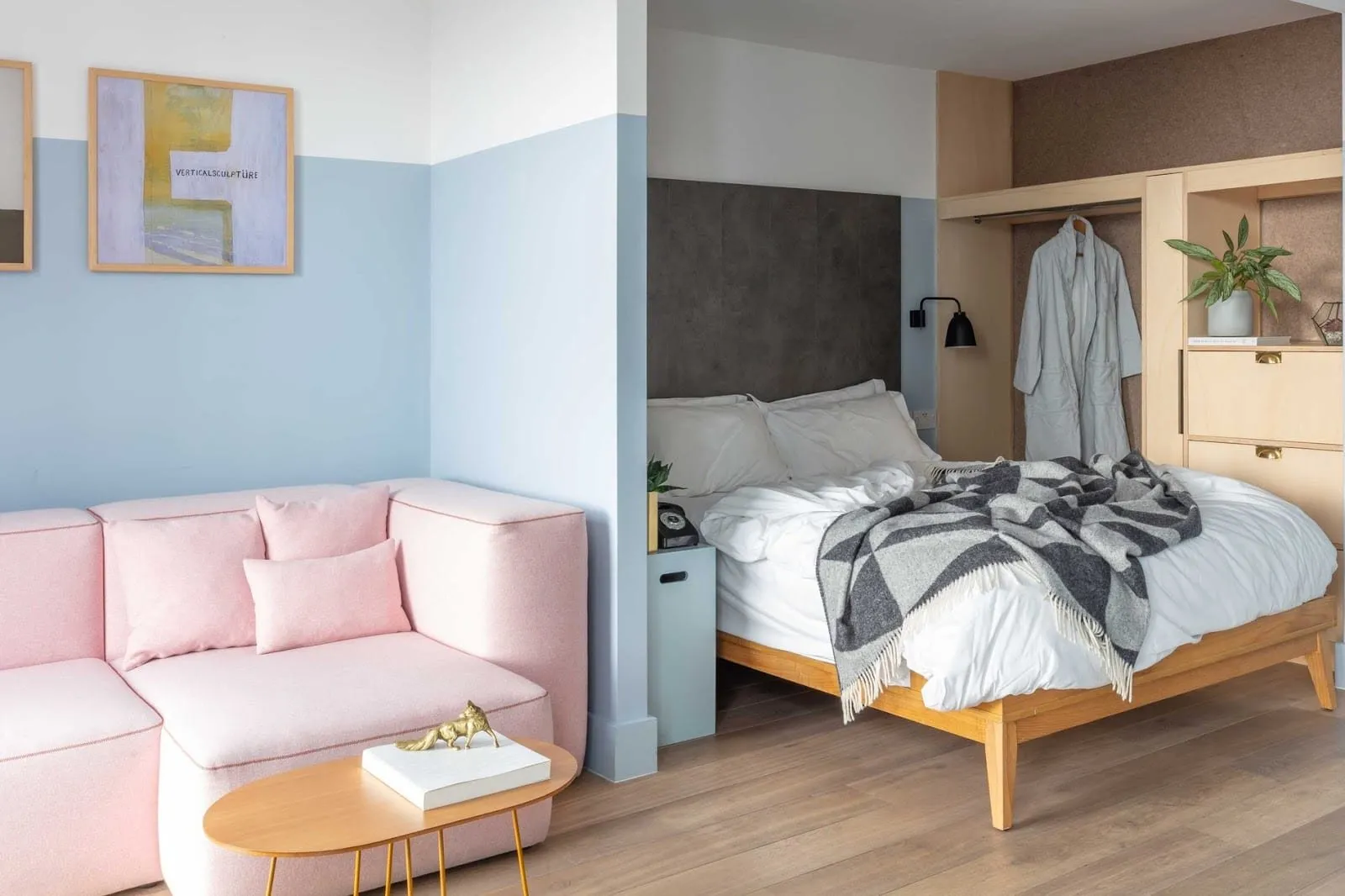 A studio at Leman Locke with a pink sofa and oval coffee table against pale blue walls, and a double bed in an alcove beside an open plywood wardrobe with a hanging robe