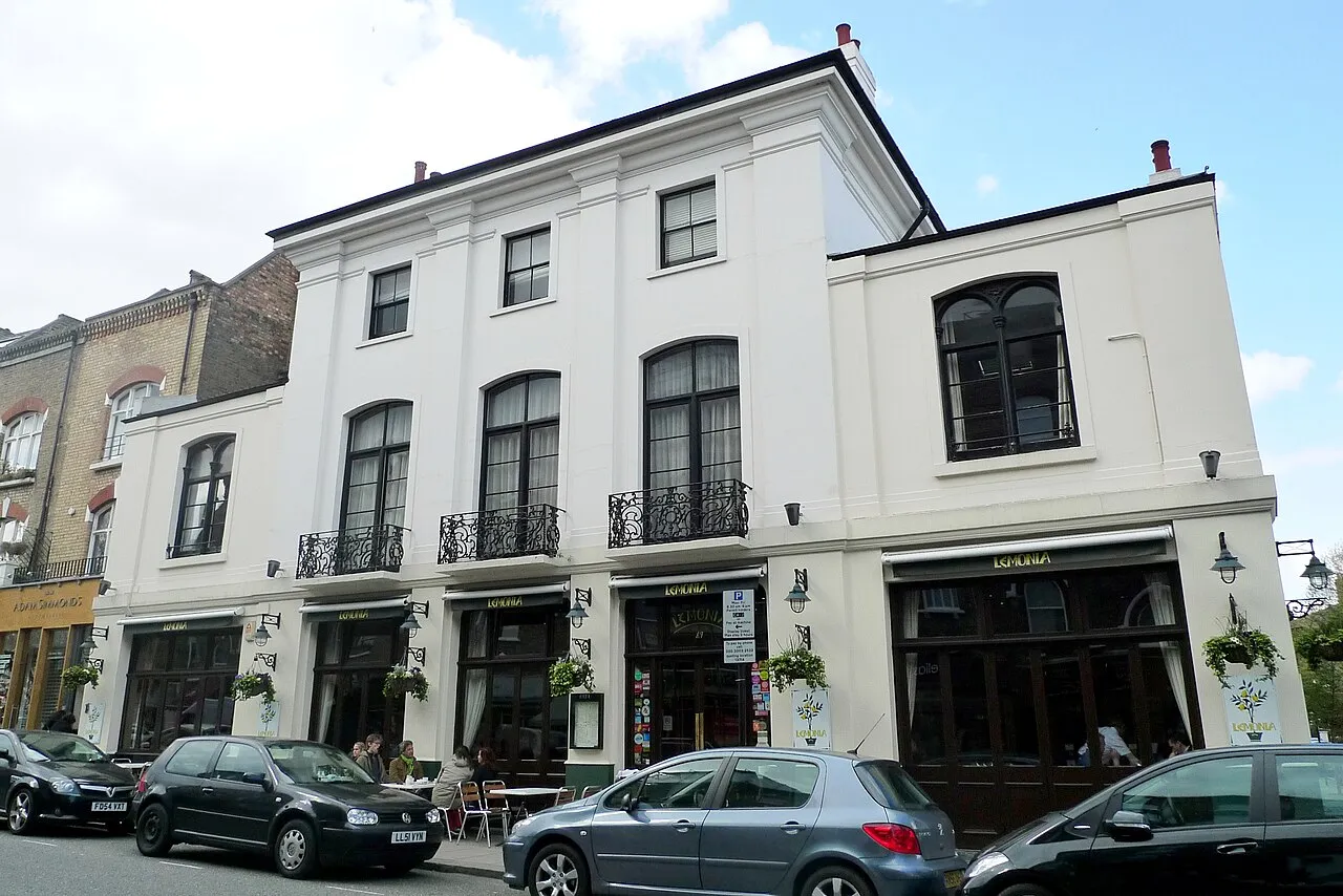 The white stucco front of Lemonia on Regent's Park Road, a former pub with tall arched windows, iron balconies and green awnings over pavement tables
