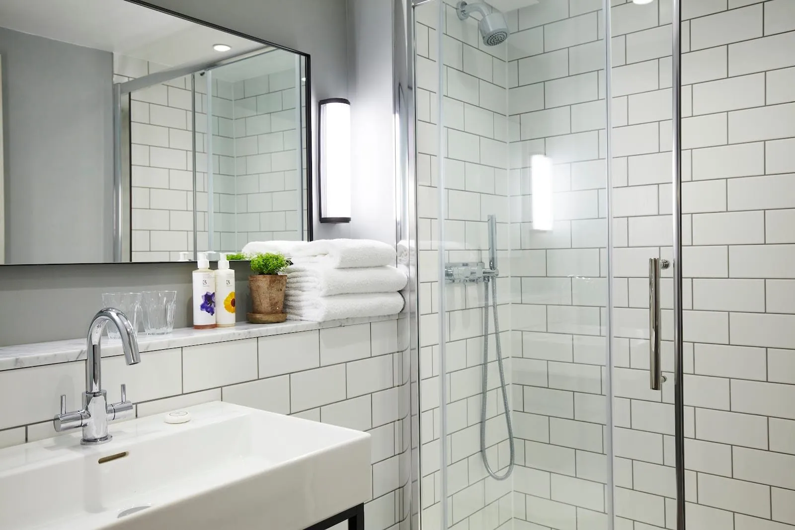 A bathroom at the Lime Tree Hotel tiled in white metro tiles, with a walk-in glass shower, a wide white basin and a stack of towels on a marble shelf