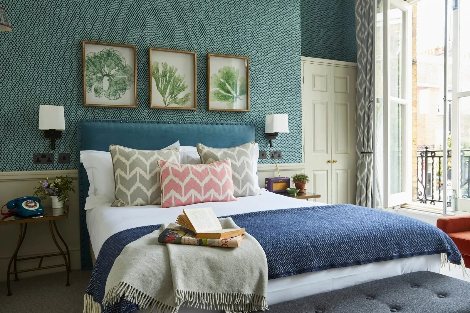 A double room at the Lime Tree Hotel with teal patterned wallpaper, a blue velvet studded headboard, three framed seaweed prints and tall French windows open onto an iron balcony over Ebury Street